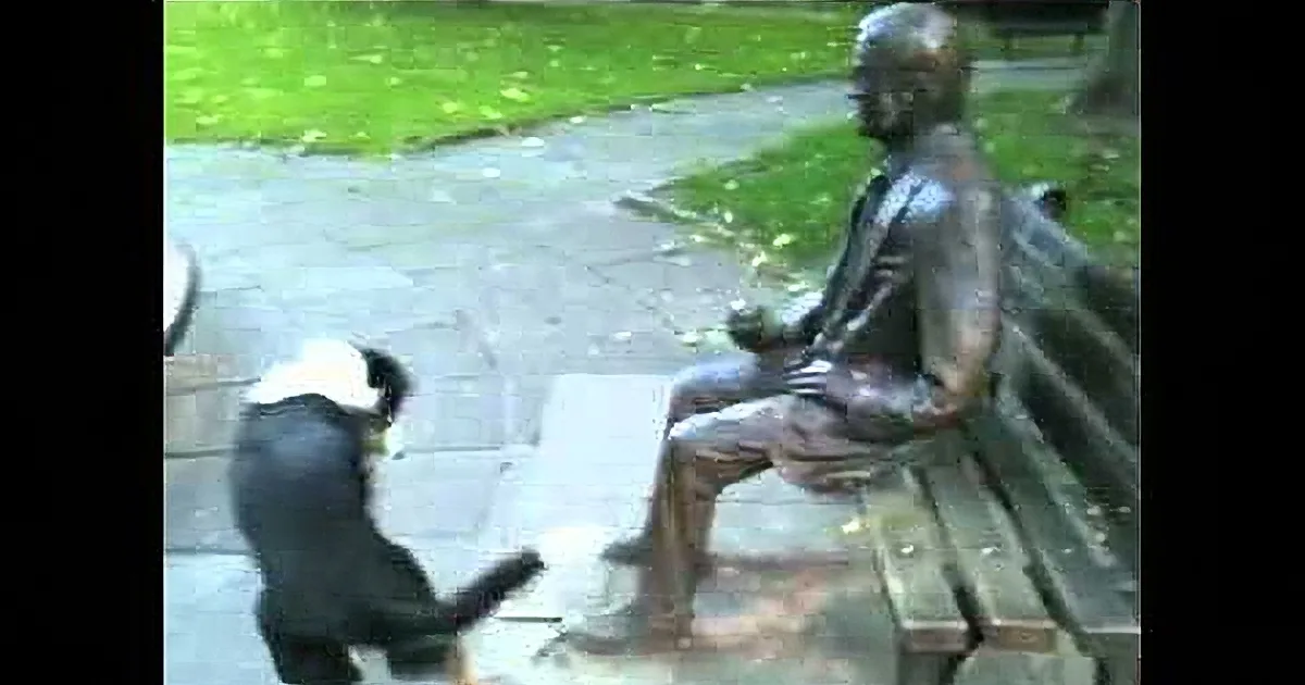 A black dog wearing a white hat interacts with a bronze statue of a seated man in a park.