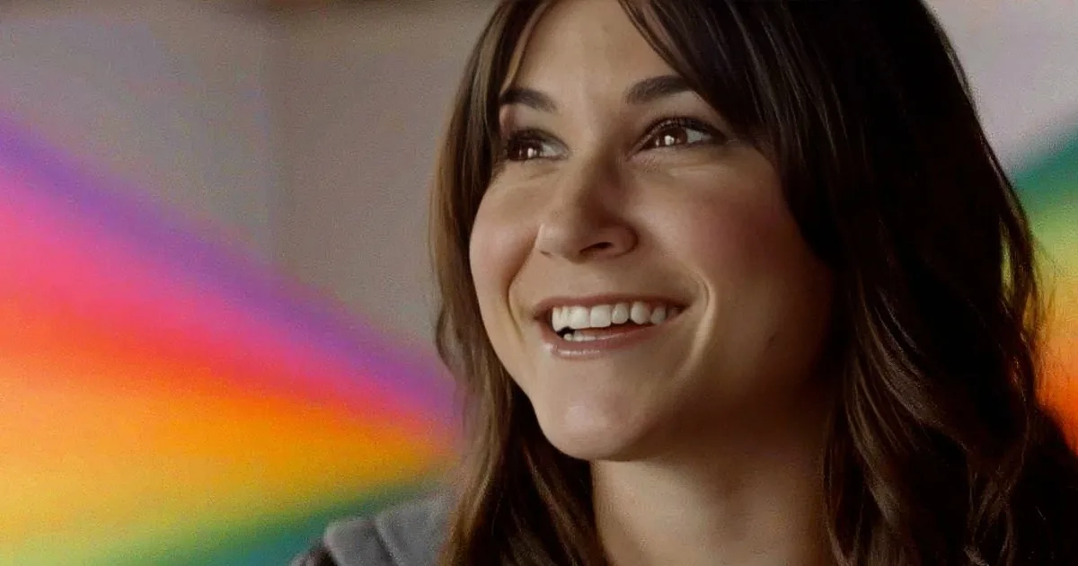 A smiling woman with long brown hair, wearing a gray top, against a colorful, blurred background.