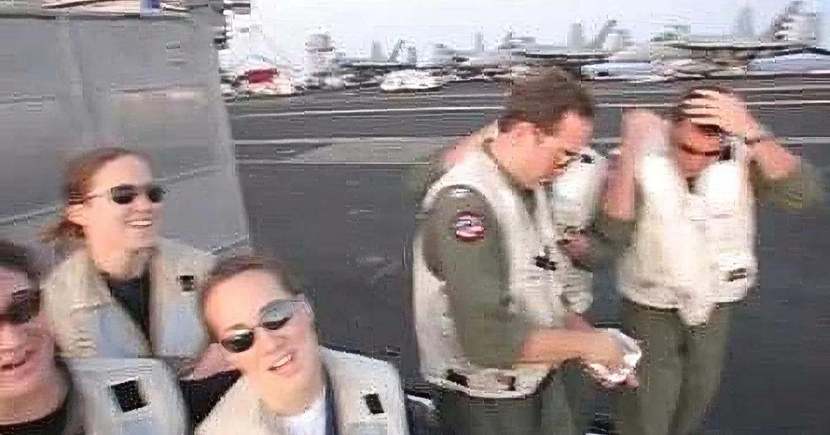 A group of people in flight suits on an aircraft carrier, with planes in the background.