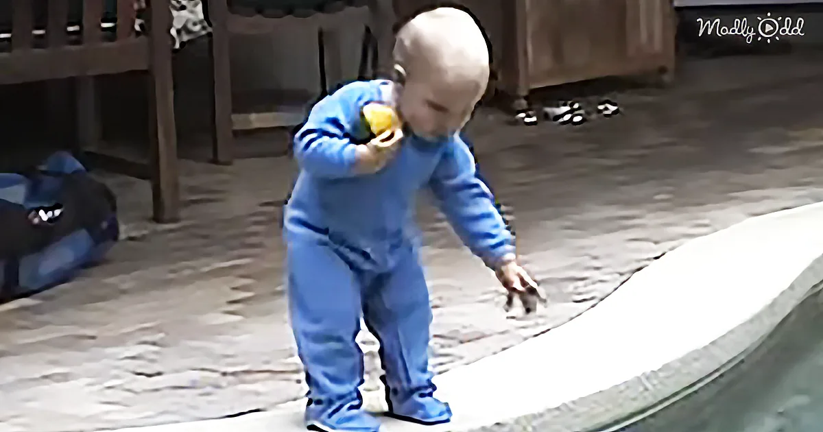 A baby in a blue onesie stands on a trampoline, holding a yellow object near its face.