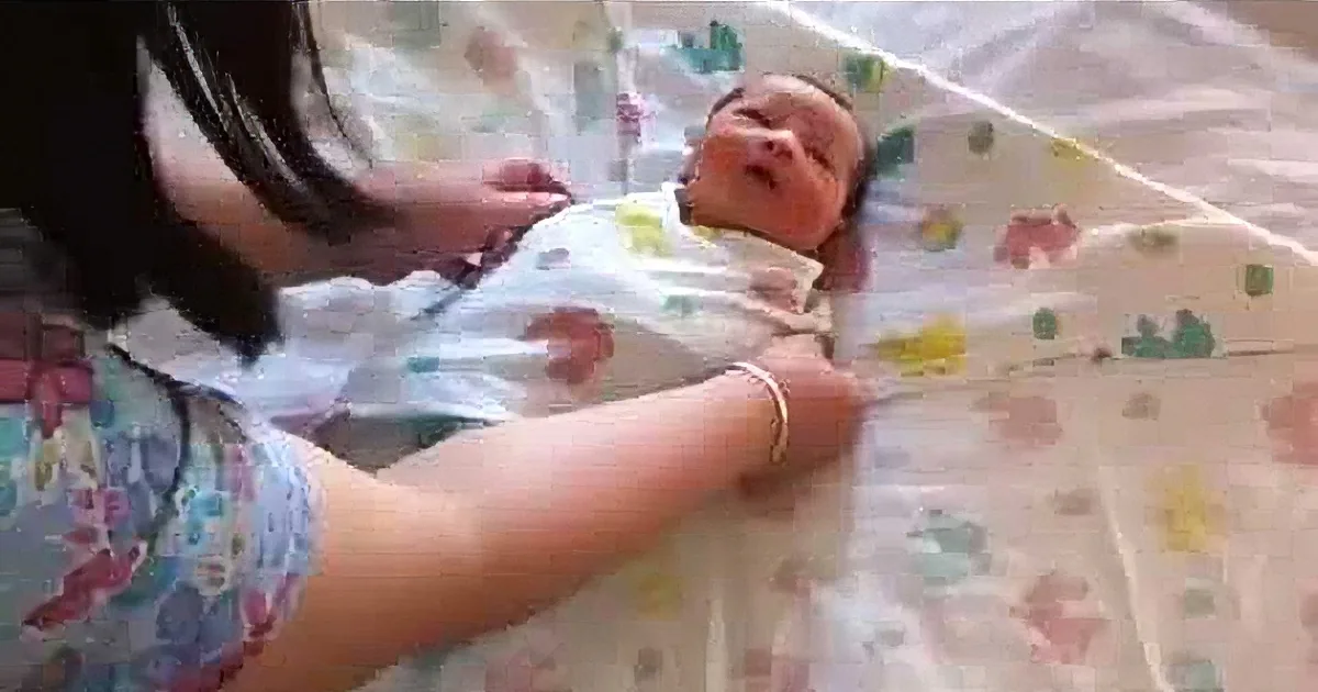 A newborn baby is swaddled in a colorful blanket, lying on a patterned surface, with a person adjusting the wrap.