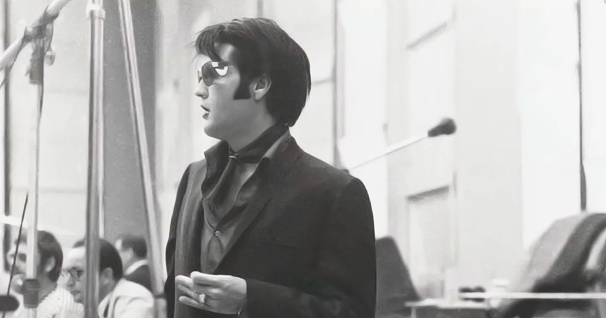 A man in a suit and sunglasses stands in a recording studio, holding a cup, with microphones and other equipment around him.
