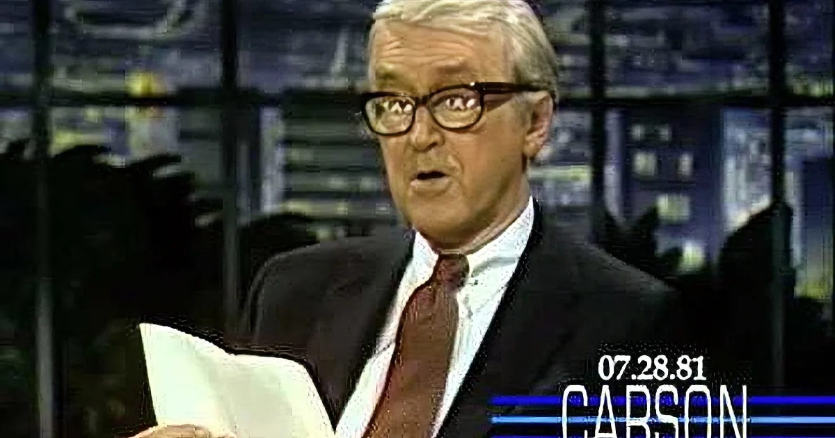 A man in a suit and glasses holds a piece of paper, appearing to speak or read.