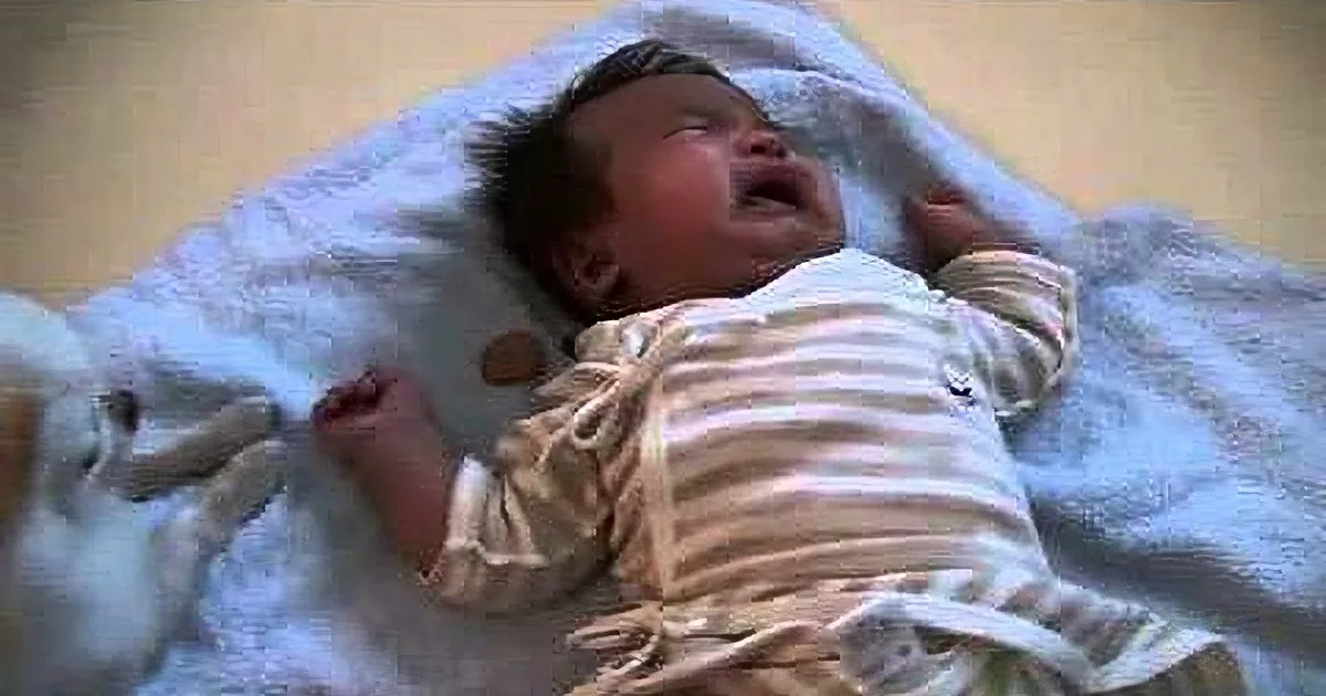 A newborn baby in striped pajamas lies on a white blanket, appearing to cry.