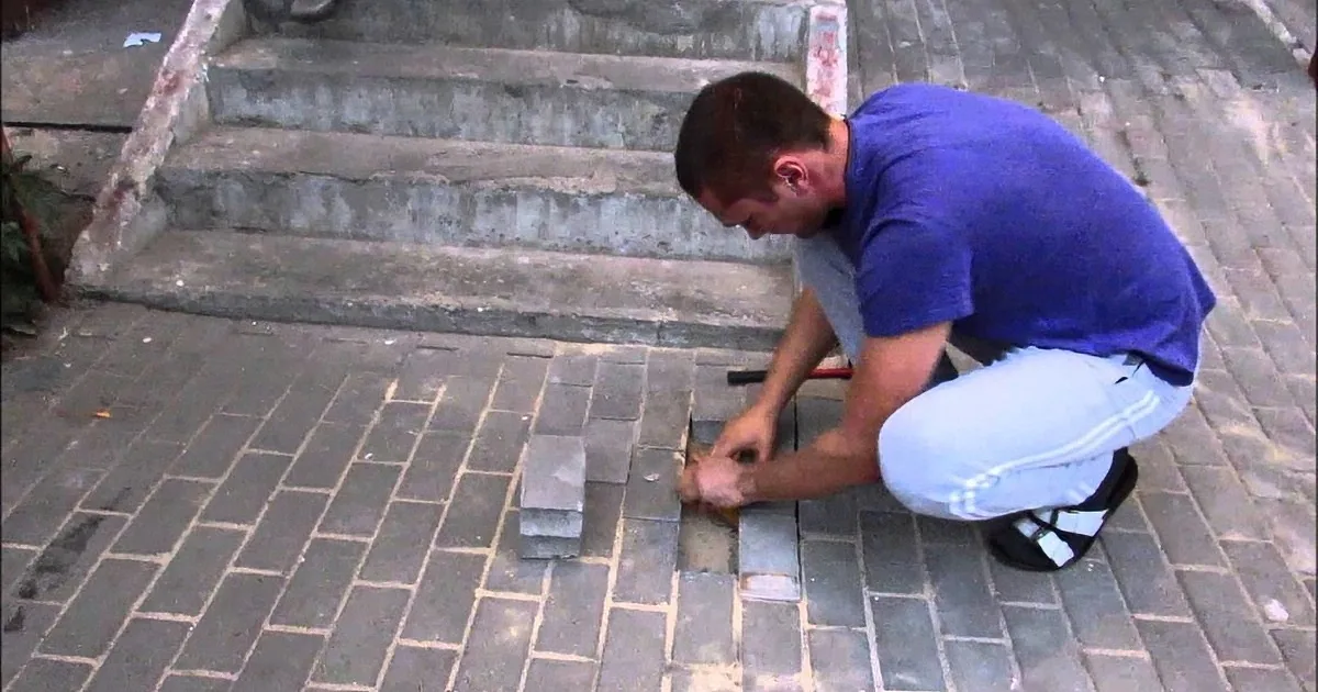 A man in a blue shirt and white pants is kneeling and working on a brick pavement, using a tool to lay bricks.