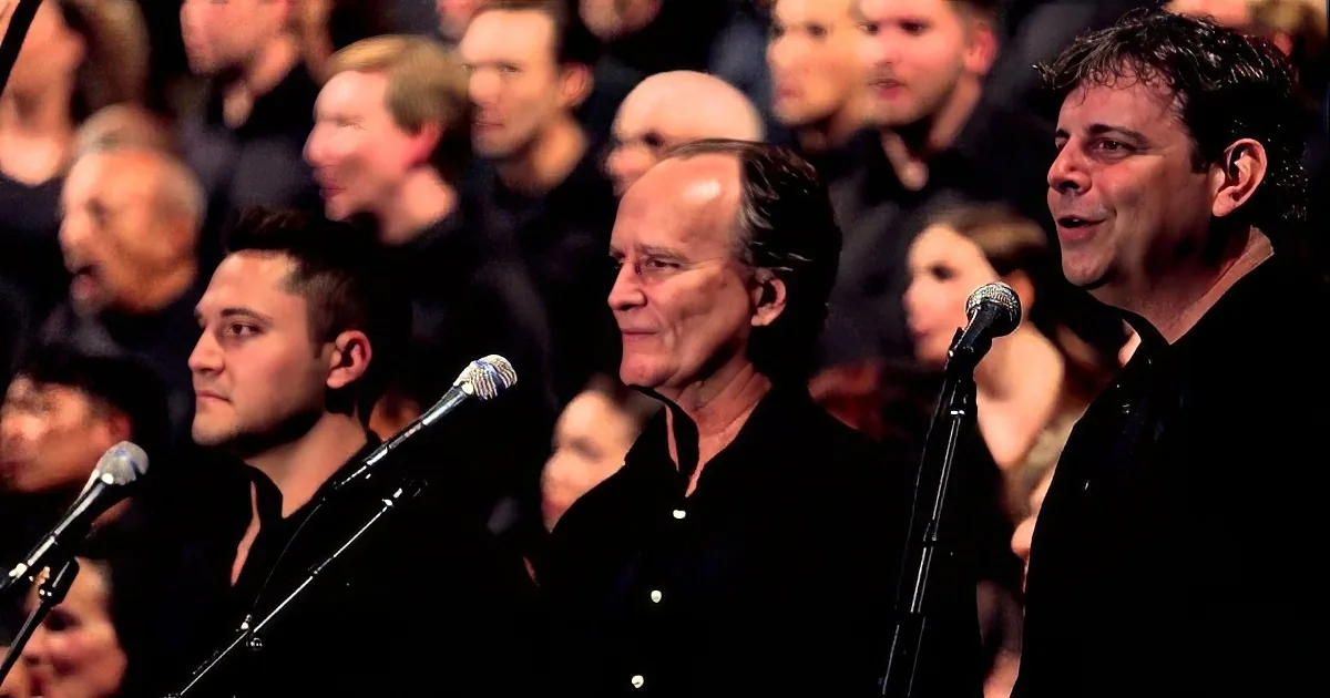 A group of men in black shirts sing into microphones in front of a blurred audience.
