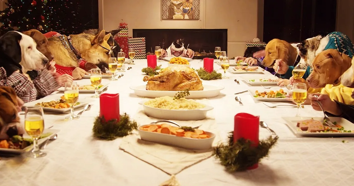 Dogs sit at a festive table with food, candles, and wine glasses, seemingly enjoying a holiday feast.
