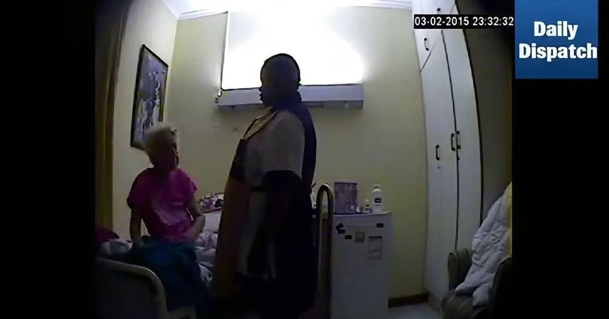 A man stands by a bed, holding a pink cloth, while an elderly woman sits nearby in a dimly lit room.