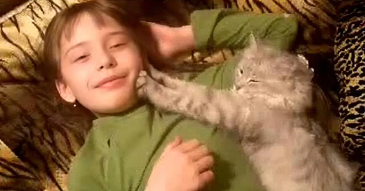 A young girl in a green shirt is lying down, smiling, with a cat on her chest.