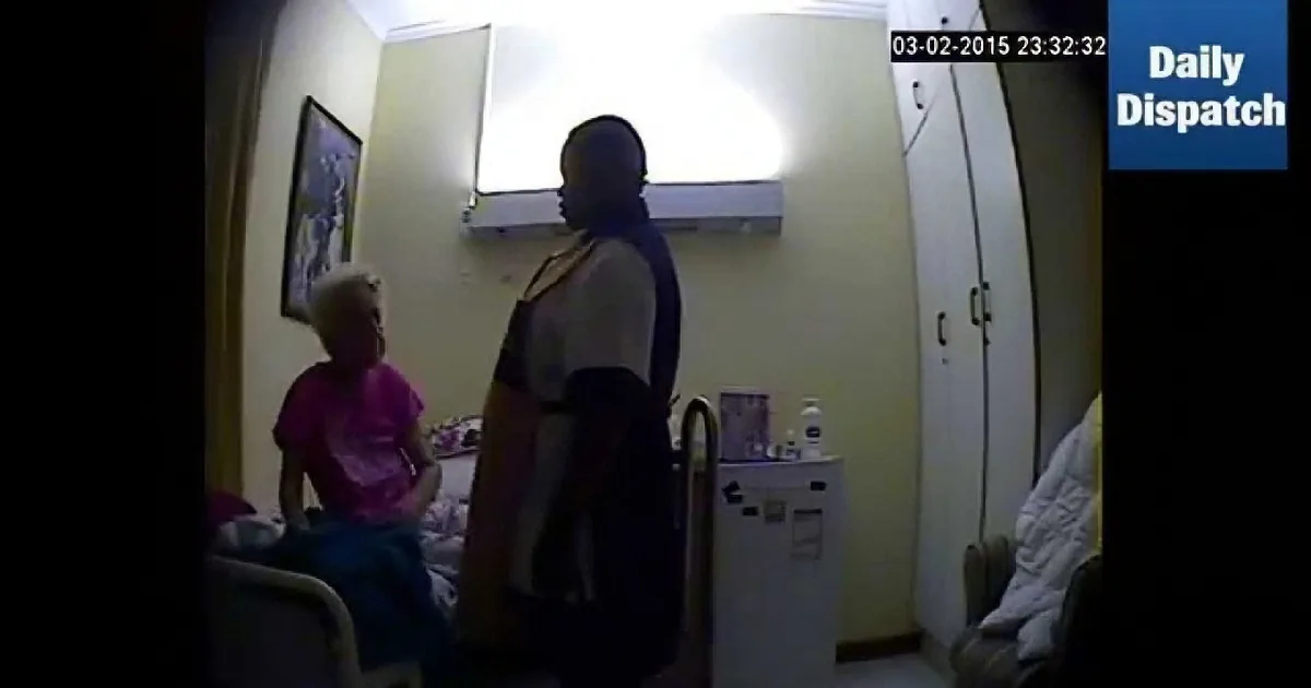A man stands near a bed, holding a pink cloth, while an elderly woman sits in a chair nearby.