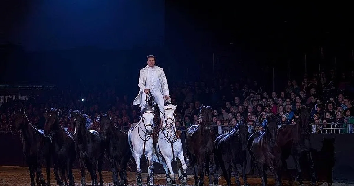 A man in white attire rides a white horse amidst a group of black horses in a dimly lit arena.