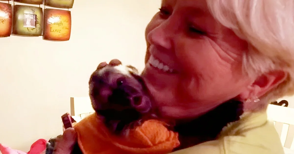 An elderly woman with white hair smiles while holding a small, dark-colored dog.