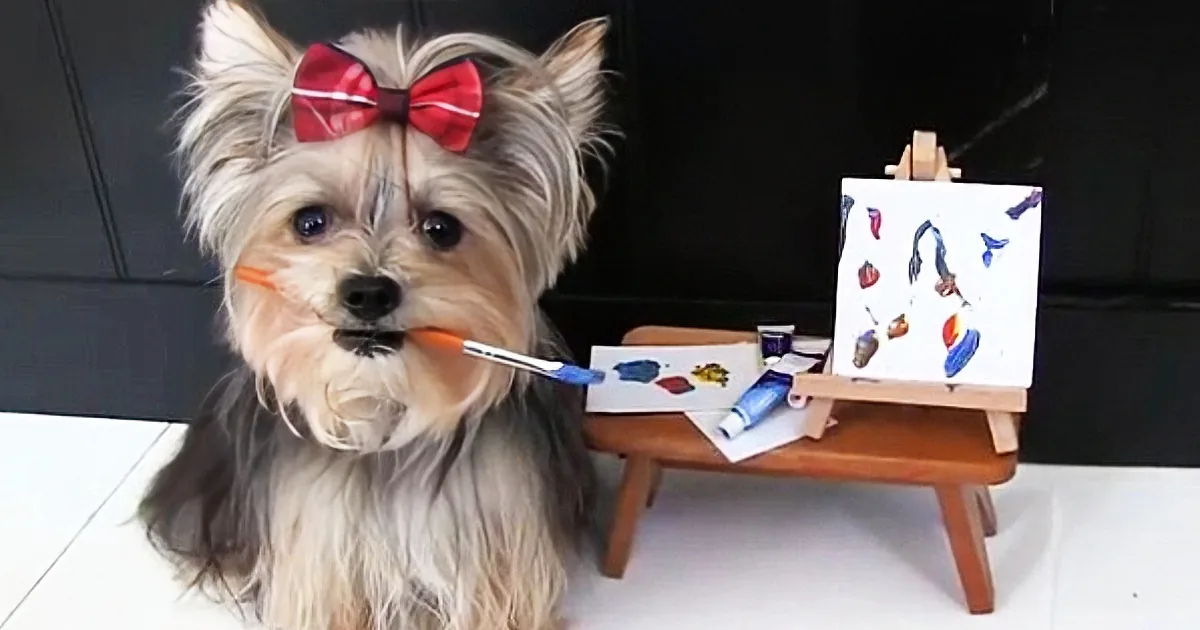 A Yorkshire Terrier with a red bow sits at a small easel, holding a paintbrush, surrounded by paint supplies.