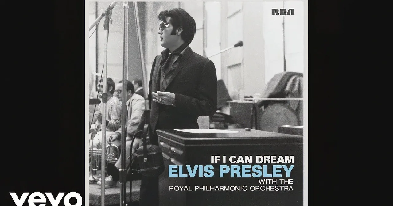 Elvis Presley stands in a recording studio, wearing a suit and sunglasses, with the Royal Philharmonic Orchestra in the...