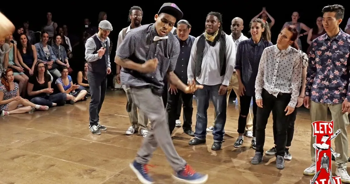 A dancer in a black and white shirt and red sneakers performs in front of a group of onlookers.