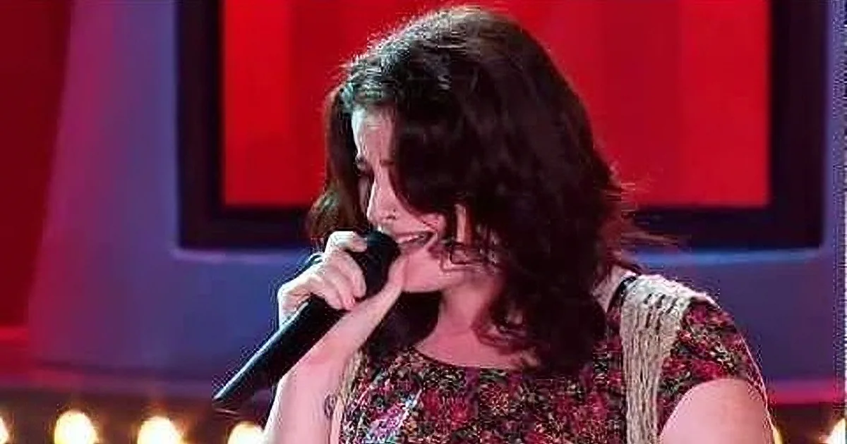 A woman with curly hair sings into a microphone on stage, wearing a floral dress.