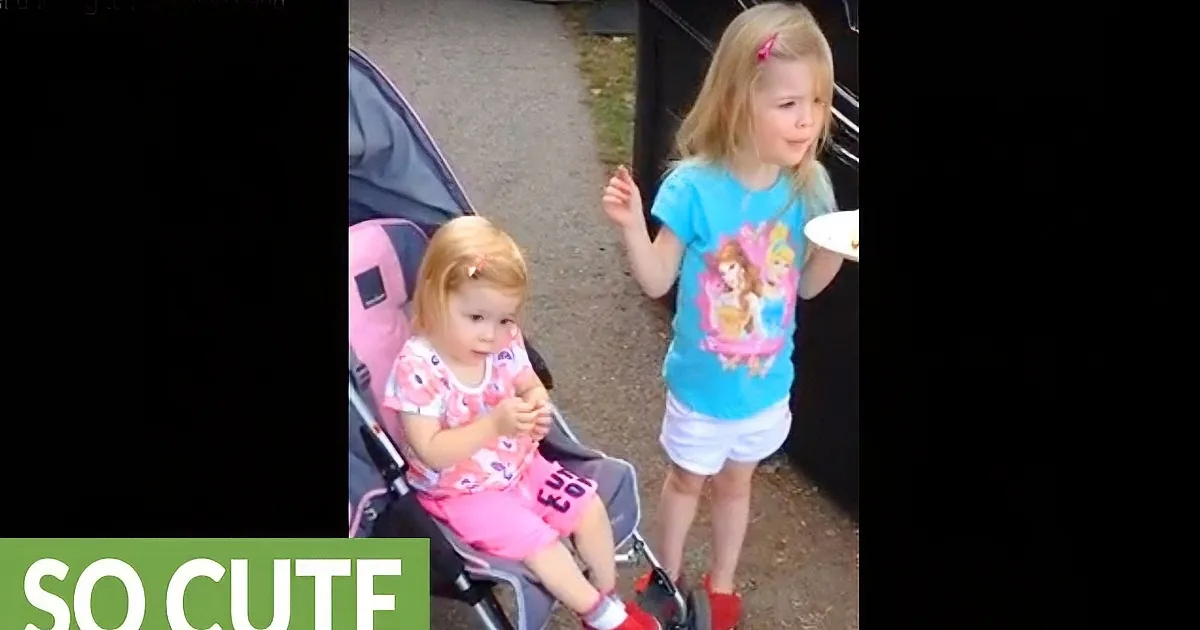 A young girl in a stroller looks at another girl standing nearby, both wearing colorful outfits.