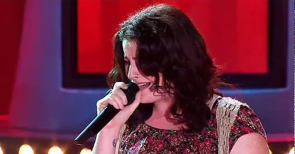 A woman with curly hair sings into a microphone, wearing a floral dress, with red and purple lighting in the background.
