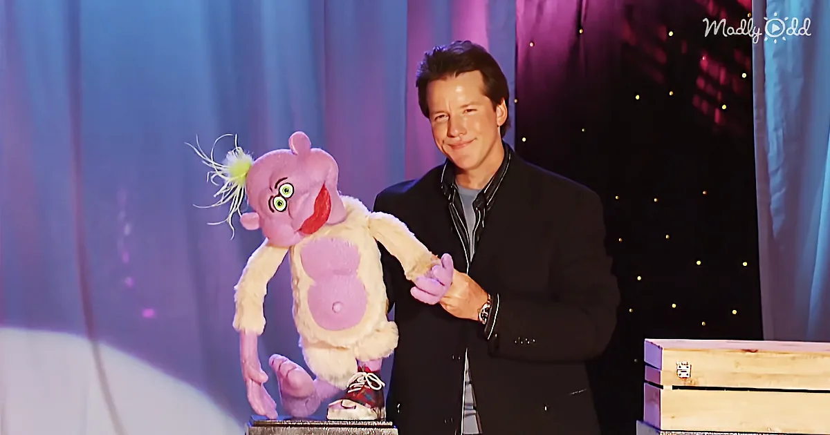 A man in a suit holds a puppet with a pink head and yellow eyes, standing on a stage with a curtain backdrop.