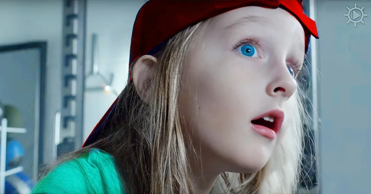 A young person wearing a red cap and green shirt looks surprised, with blue eyes and long blonde hair.