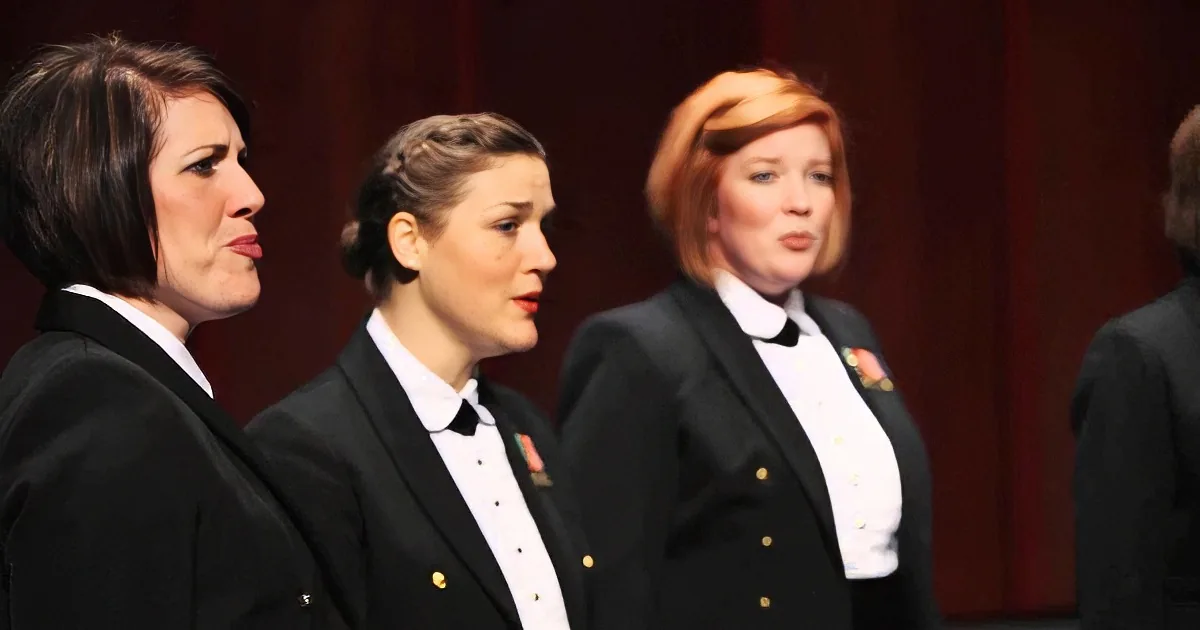 Three women in formal attire sing in a choir, with one woman in the foreground.