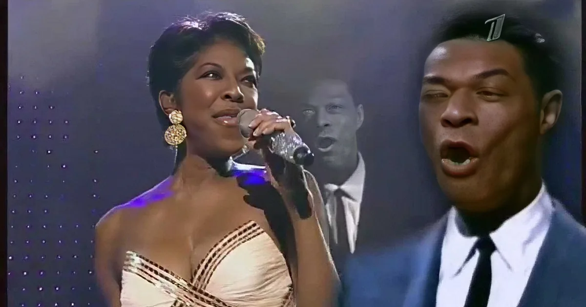 A woman in a white dress sings into a microphone, with a man in a suit singing beside her.