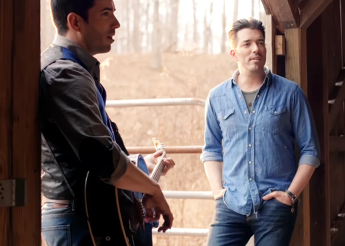 Two men, one holding a guitar, standing in a rustic setting, engaged in conversation.