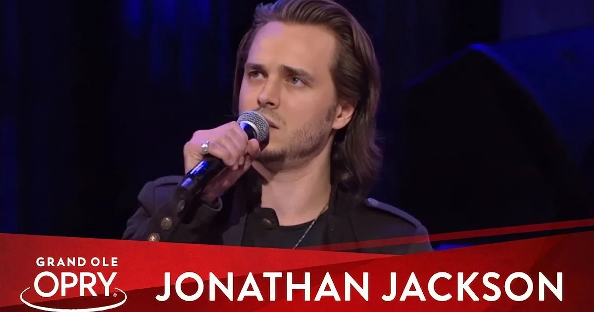 Jonathan Jackson sings at the Grand Ole Opry, holding a microphone with a red banner below.