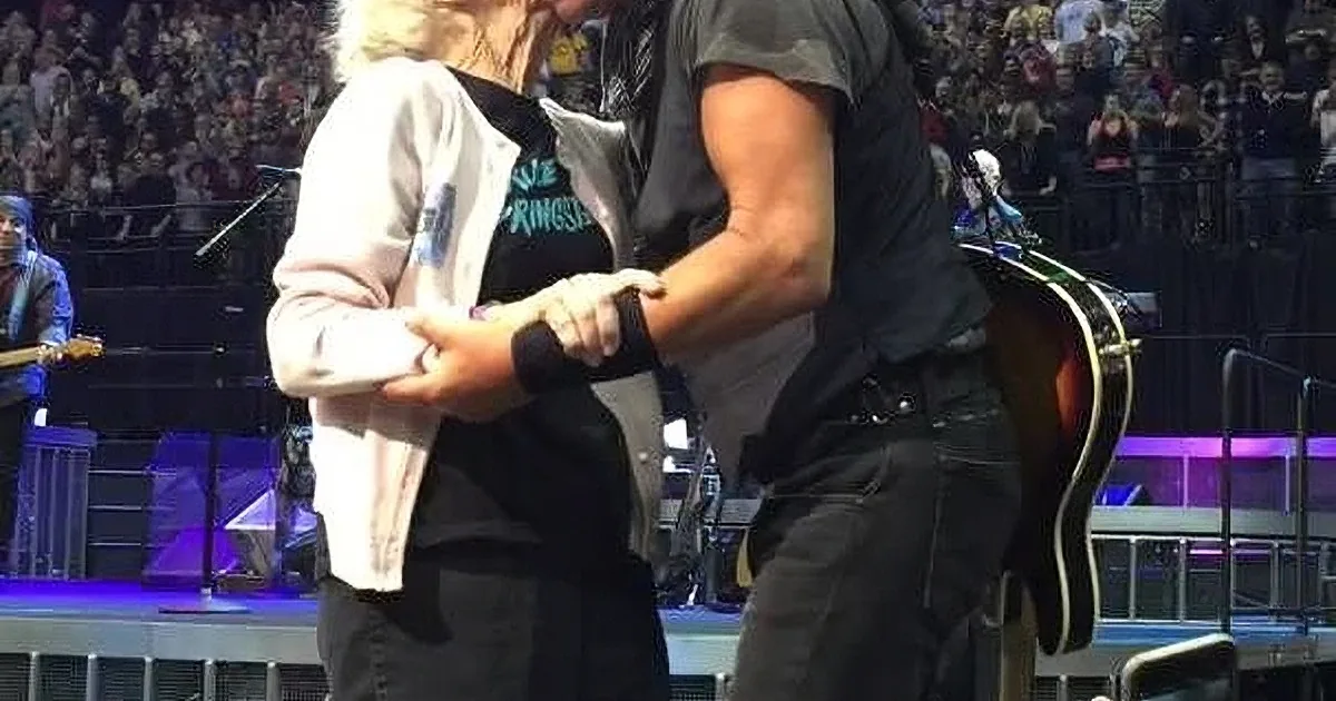 A man and woman are dancing on stage, surrounded by a large audience, with a guitarist in the background.