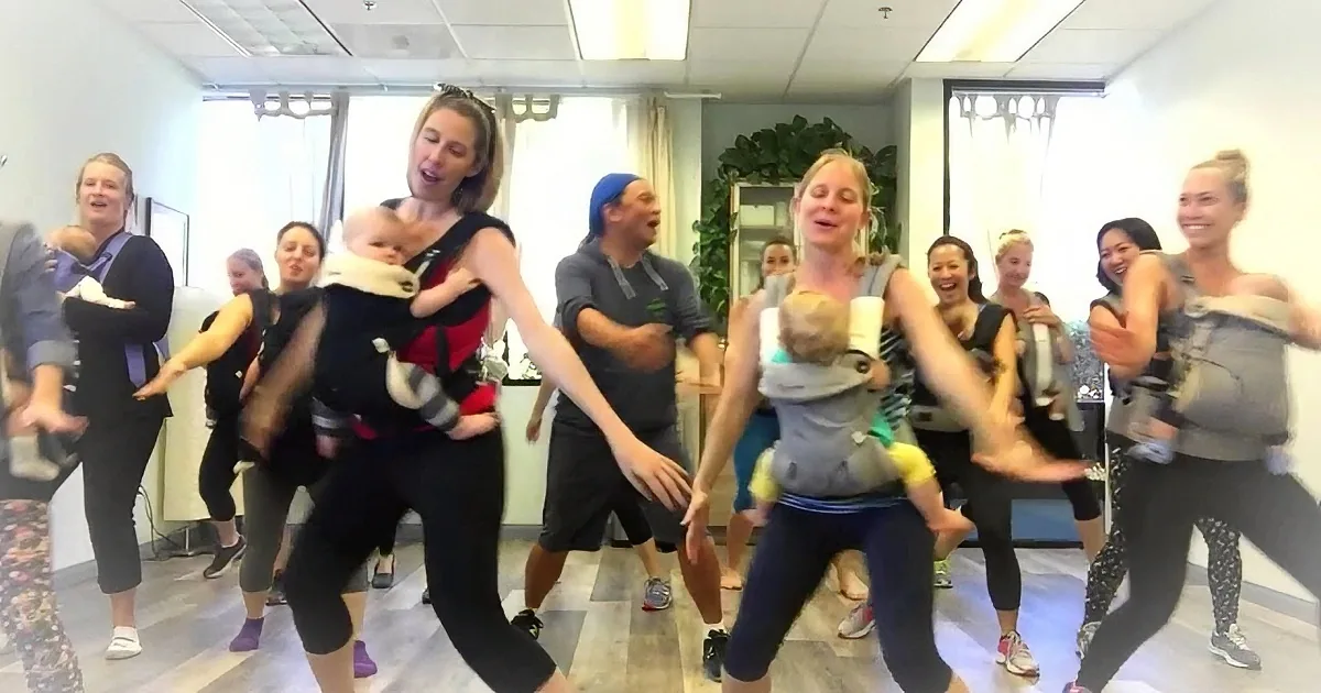 A group of people, including women with babies in carriers, are dancing in a studio.