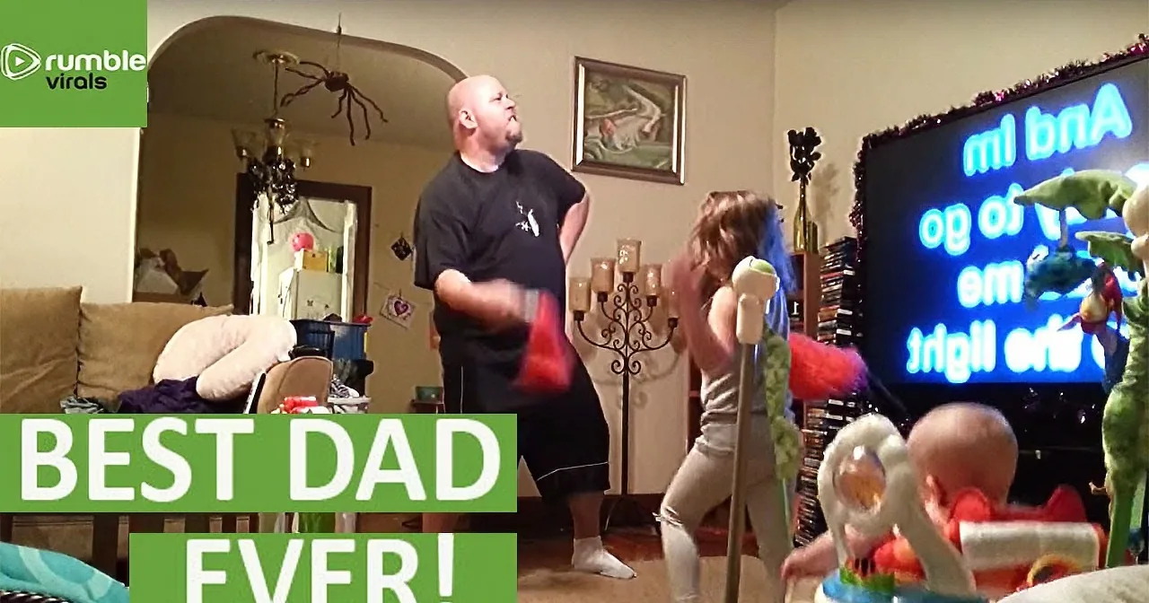 A man dances joyfully in a living room with a baby and a TV showing a puppet show.