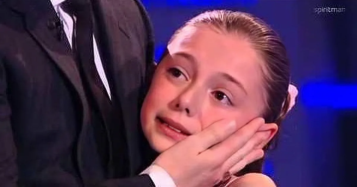 A young girl with a surprised expression, hands covering her mouth, in a dimly lit setting.