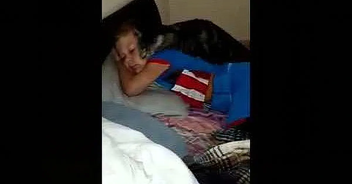 A person is lying on a bed, partially covered by a blanket, with a blue object and a red and white striped item nearby.