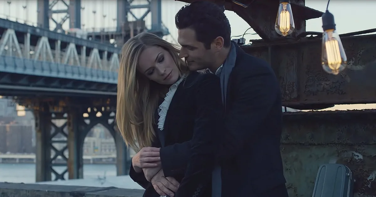 A couple embraces warmly, standing by a bridge with hanging lights, suggesting a romantic moment.