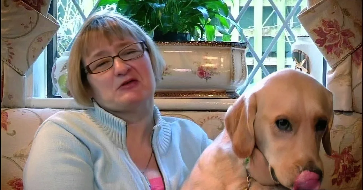 A woman with glasses sits on a floral-patterned couch next to a dog licking its nose.