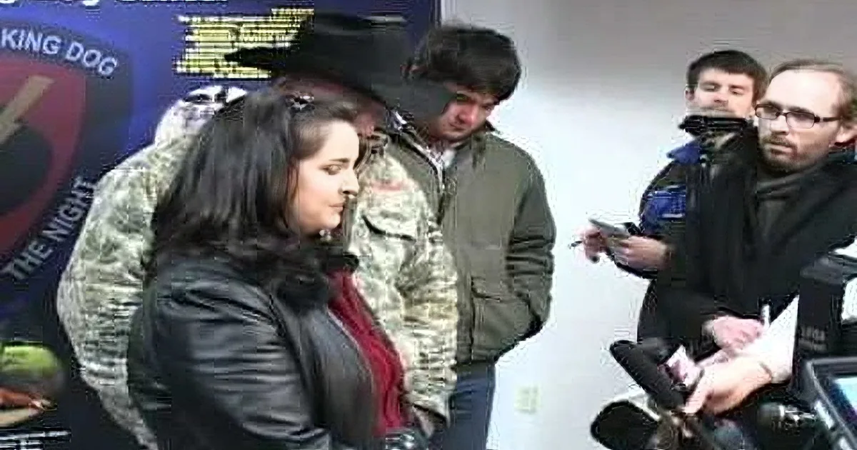 A woman in a black jacket stands in front of a poster, surrounded by people and microphones.