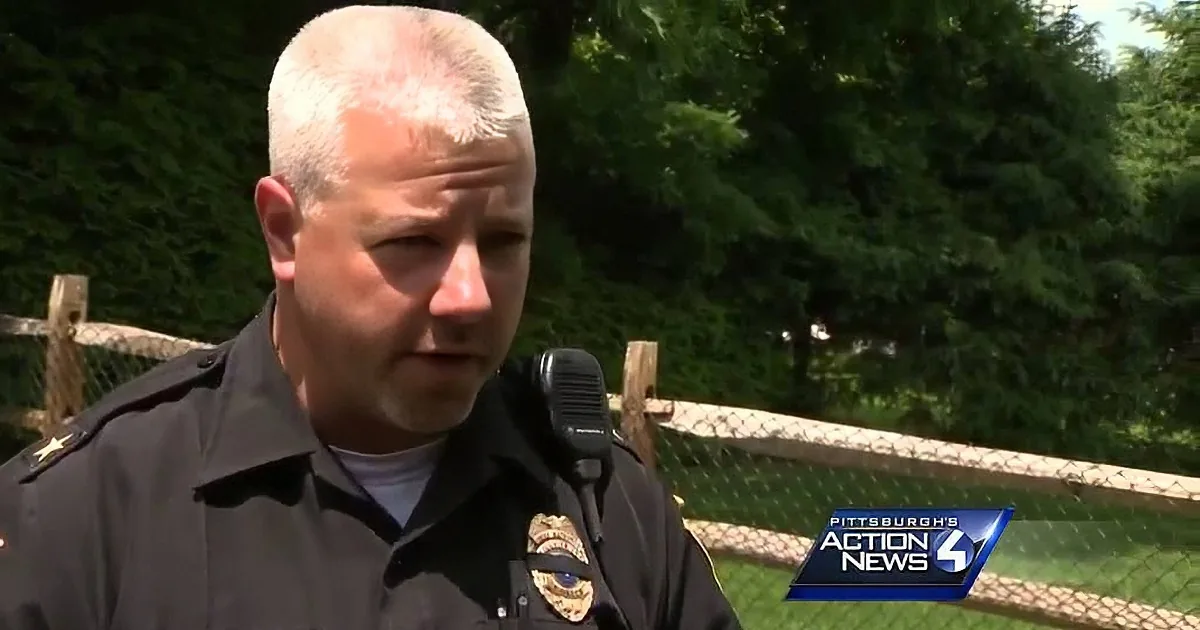 A police officer in uniform stands outdoors, with a chain-link fence and trees in the background.