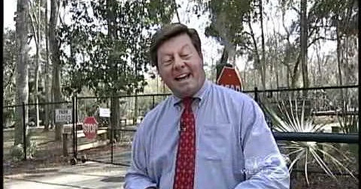 A man in a blue shirt and red tie stands outdoors, holding a glass, with trees and a fence in the background.