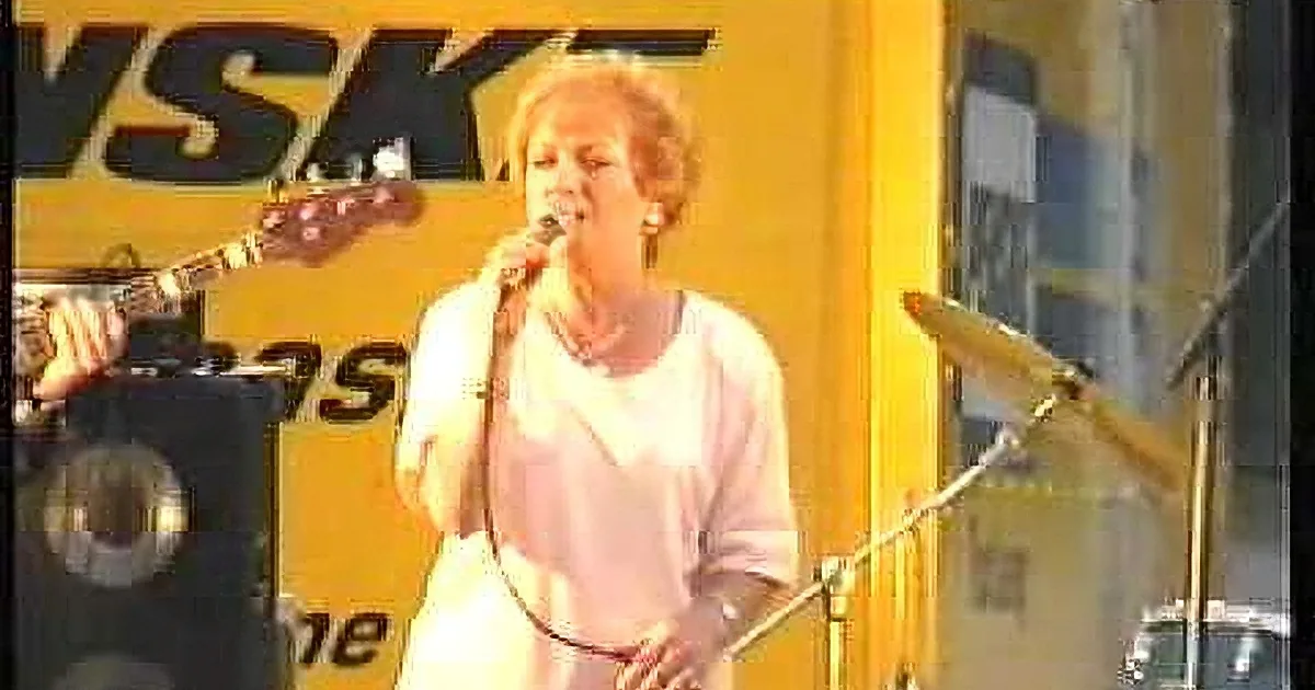 A woman sings into a microphone on stage, surrounded by musical equipment and a yellow background.