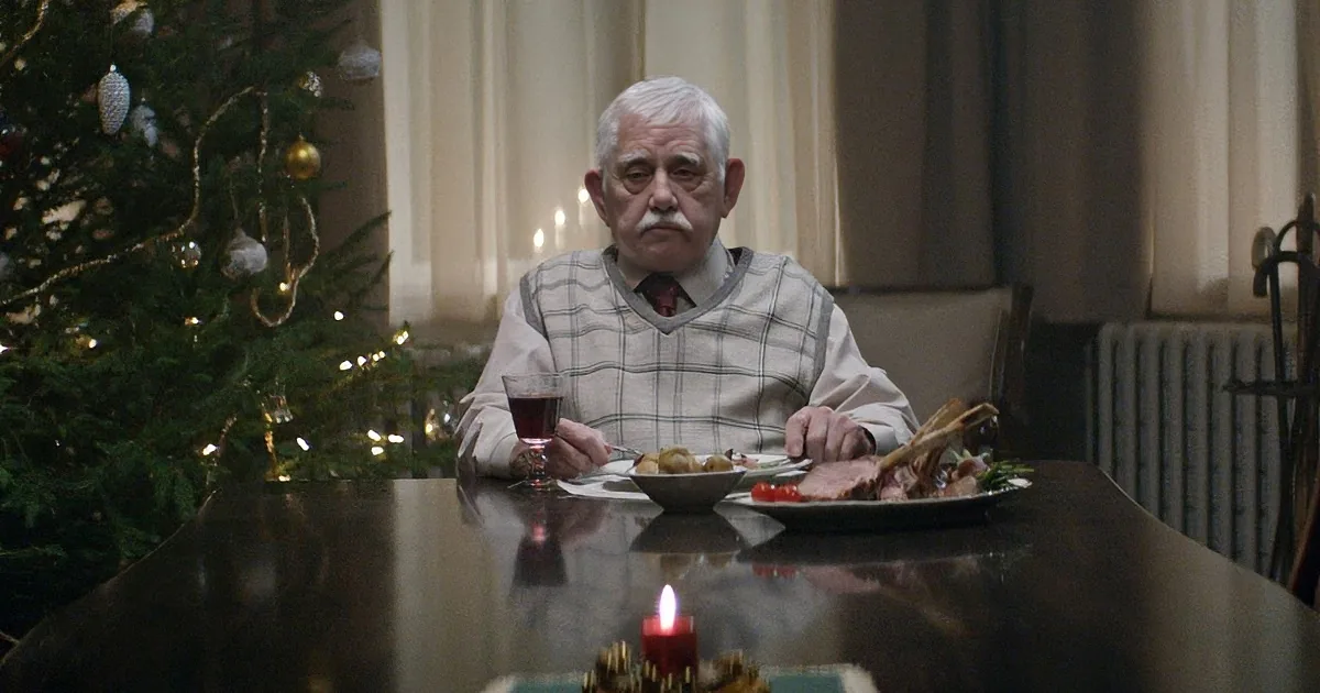 An elderly man sits at a table with a Christmas tree in the background, surrounded by holiday decorations and food.