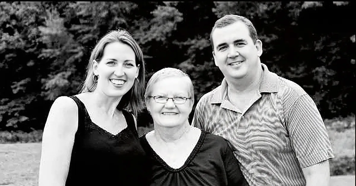 A woman in a black dress smiles with two others in a black-and-white photo.