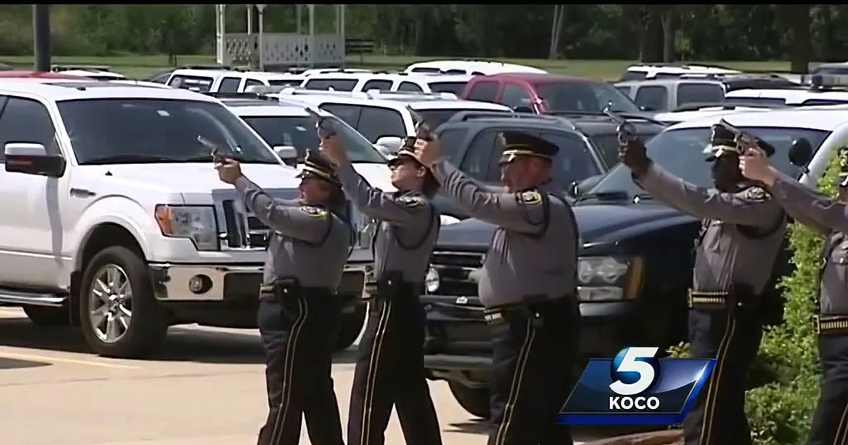 Police officers in uniform fire ceremonial shots in a parking lot.