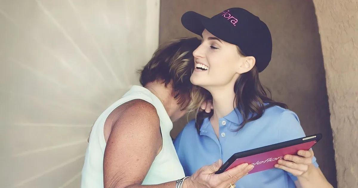A woman in a blue shirt and black cap is smiling while holding a tablet, interacting with another woman in a white top.
