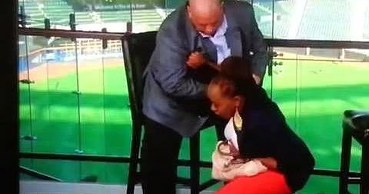 A man in a suit is shaving another man's head in a stadium setting.