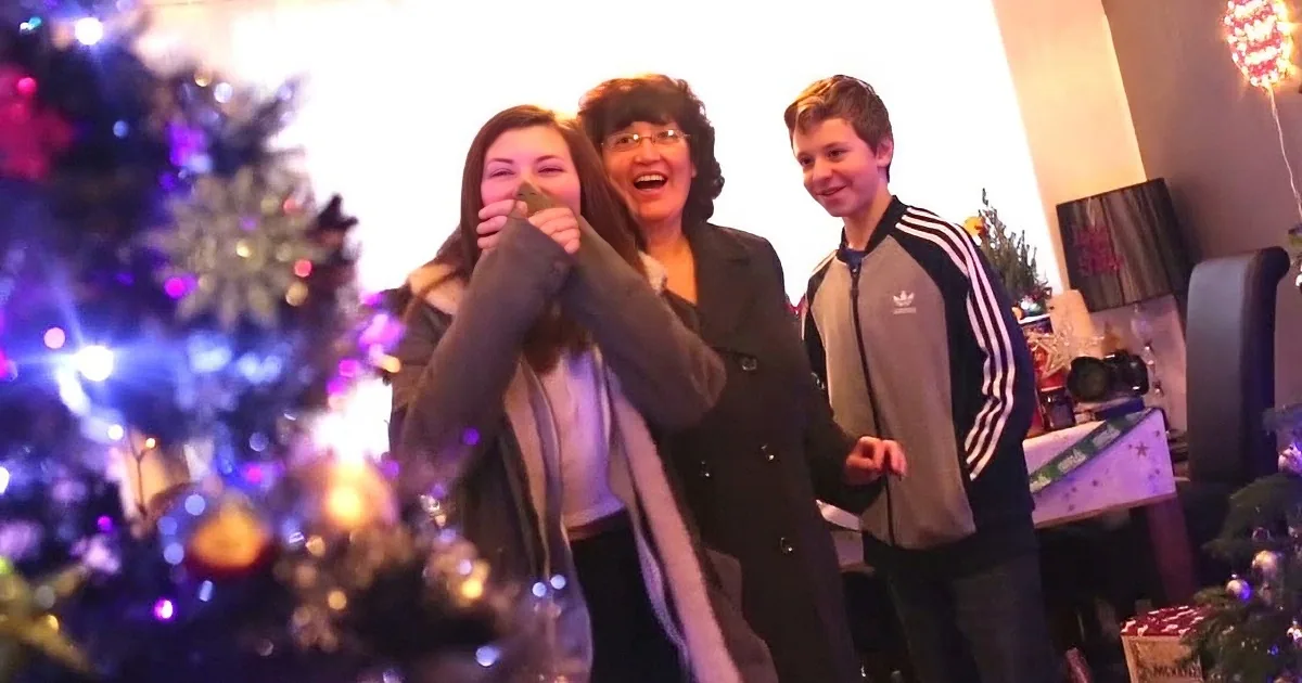A woman holds a wrapped gift, smiling with two others near a Christmas tree and presents.