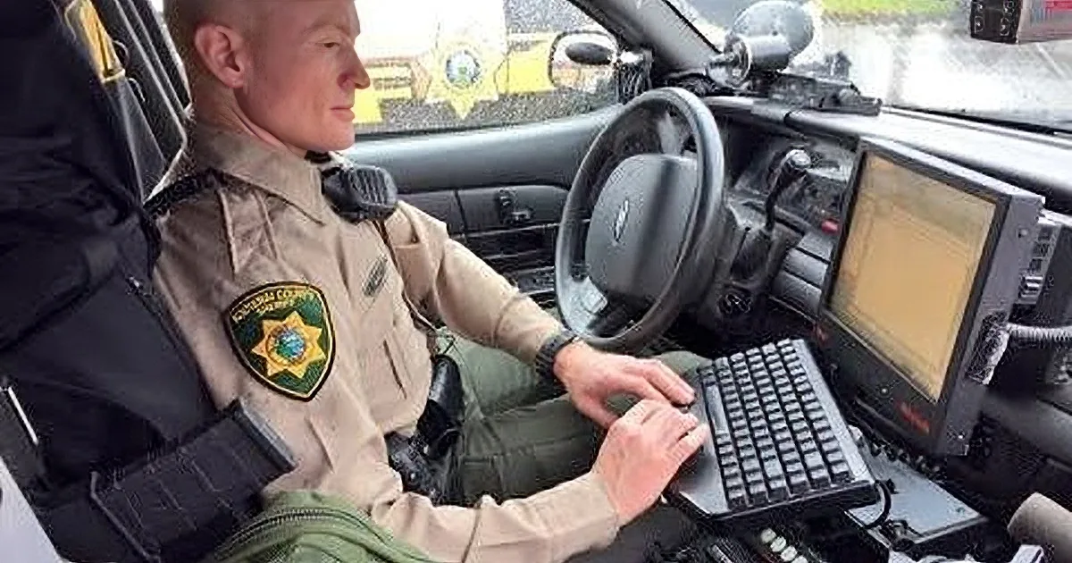 A police officer in uniform sits in a patrol car, holding a folder and looking to the side.