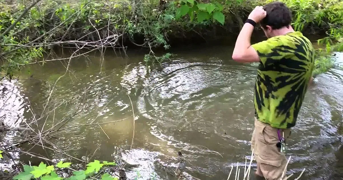 A man in a green and black shirt stands in a shallow stream, holding his head, with a branch partially submerged in the...