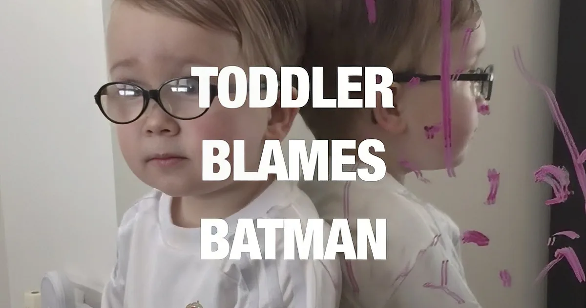 A toddler wearing glasses and a white shirt with a crown emblem is looking to the side.