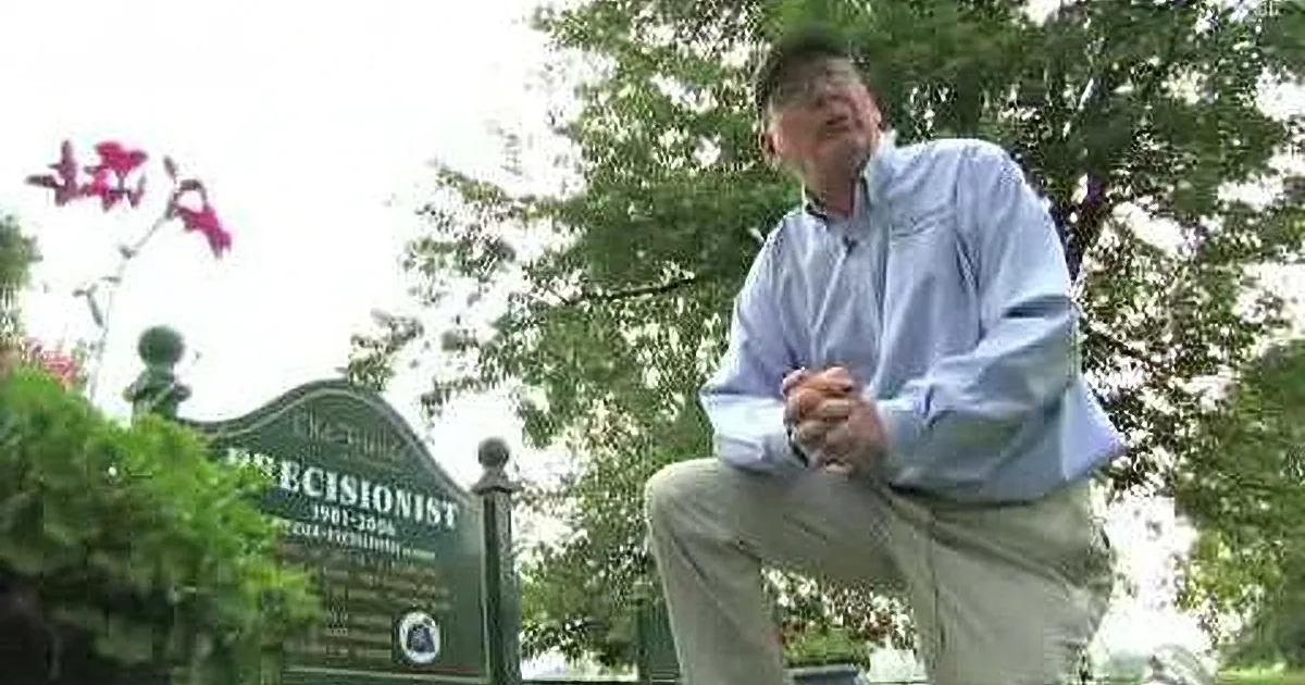 A man in a light blue shirt sits on a bench, hands clasped, in front of a sign that reads "Precisionist 1901-2006."