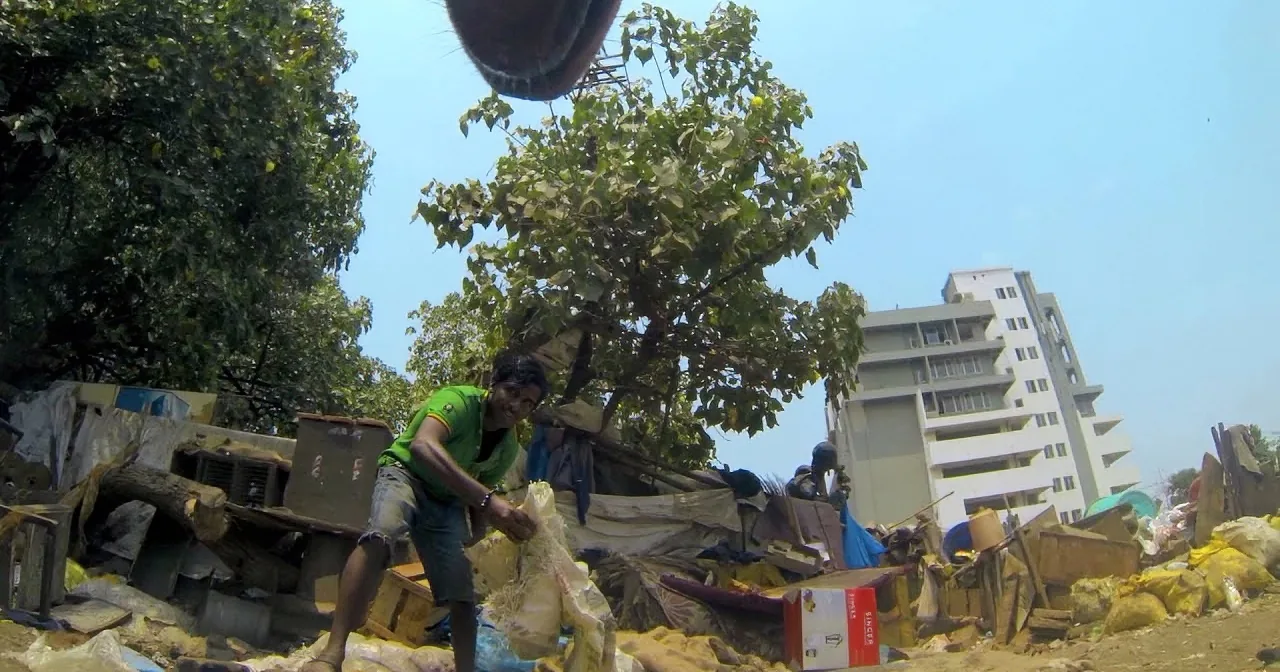 A man sorts through debris in a cluttered area with trees and buildings in the background.